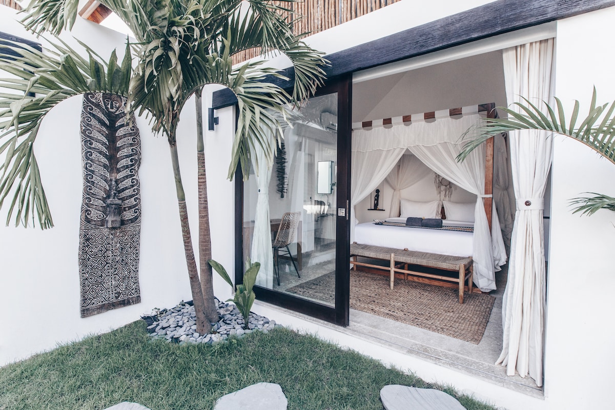 An outdoor view captures a serene bungalow surrounded by lush greenery. The spacious bedroom features a canopied king bed with a natural wood frame, complemented by soft textiles. Large glass doors invite sunlight and provide an unobstructed view of the tranquil garden area.