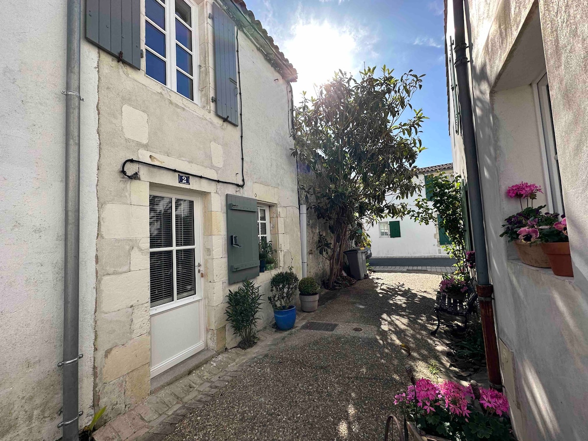 A charming entrance to a small, stone house is depicted, featuring green shutters and a welcoming door. Lush plants line the walkway, with vibrant flowers arranged in pots. Soft sunlight filters through the trees, casting a warm glow on the cobblestone path.