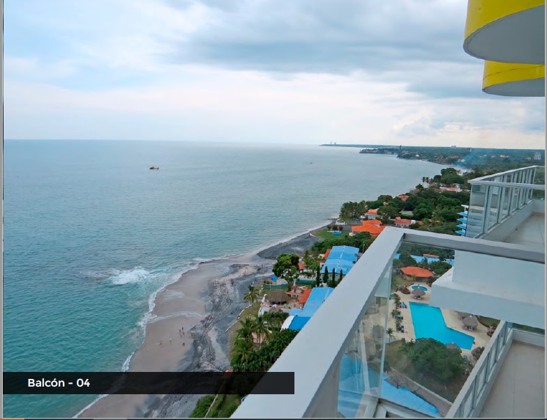 An expansive view of the ocean is captured from the balcony, featuring a blend of sandy beach and rocky shoreline. Vibrant umbrellas and lounge areas are visible below, alongside the calm blue water stretching toward the horizon under a partially cloudy sky.