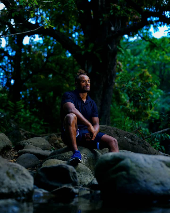 A man with a curly top knot wearing blue clothes sits on rocks and gazes contemplatively in a lush rainforest.