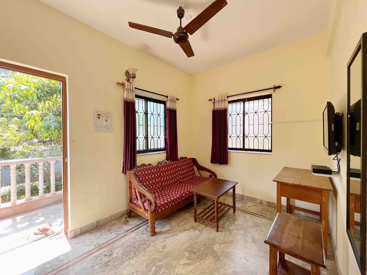 A bright living room is furnished with a wooden sofa and a simple coffee table. Two large windows allow natural light to fill the space, while dark curtains provide a contrast. A wall-mounted television is visible, enhancing functionality.