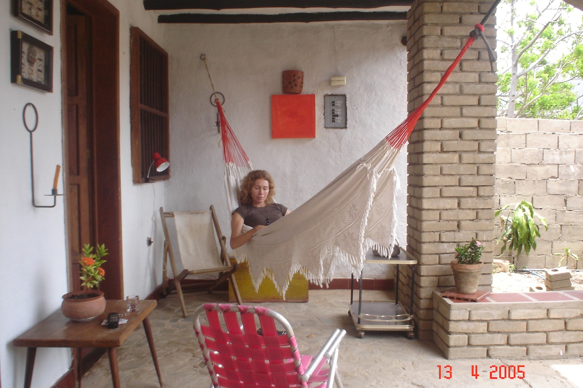 A shaded porch area features a comfortable hammock suspended between two posts, alongside a small chair and a potted plant. The textured walls and wooden accents create a cozy setting for relaxation.