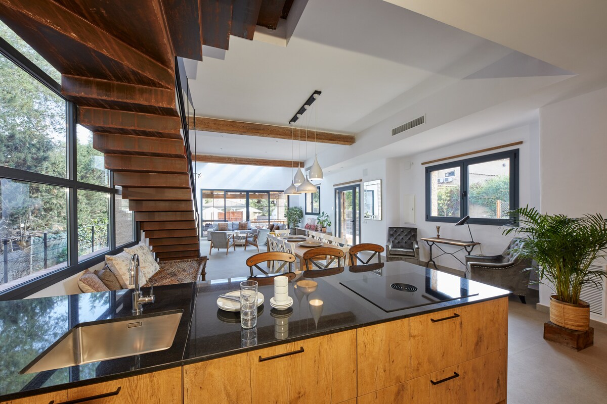 An open plan living area is highlighted, featuring a wooden staircase leading to the upper level. Large windows fill the space with natural light, while a modern kitchen with a dark countertop is visible in the foreground. Comfortable seating areas and greenery can be seen beyond.