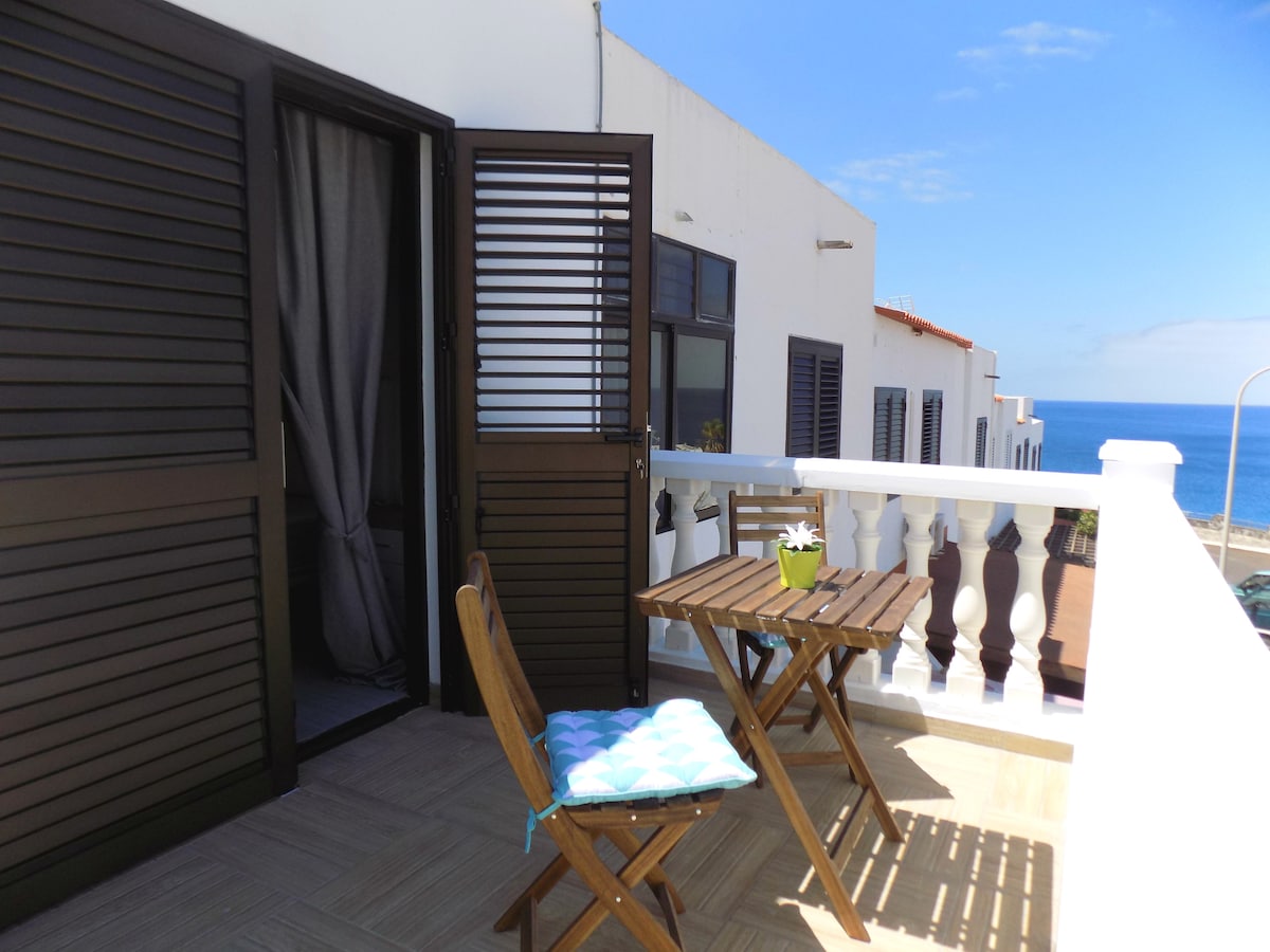 A balcony is shown, featuring a wooden table and a folding chair. A small potted plant adds a touch of greenery. The view opens up to the ocean and clear blue sky, with light reflecting off the water.