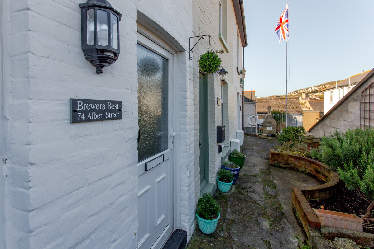 The entrance of the cottage is visible, featuring a white door with glass panels and a lantern light. Potted plants adorn the entrance, and a sign reads 'Brewers Rest, 74 Albert Street.' A Union Jack flag is seen in the background against a clear sky.
