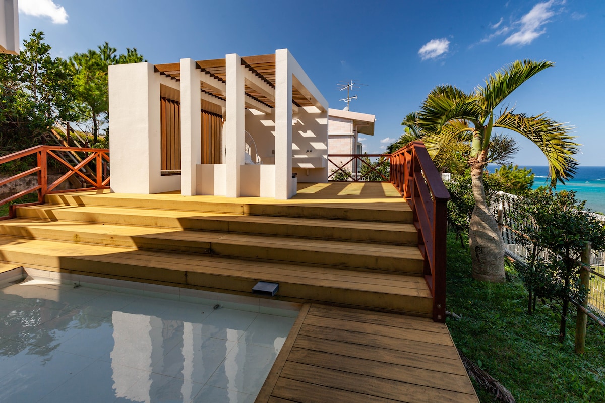 An outdoor deck is visible, featuring steps leading up to a bright, open structure with wooden accents. Lush greenery surrounds the area, and the blue ocean can be seen in the distance under a partly cloudy sky.