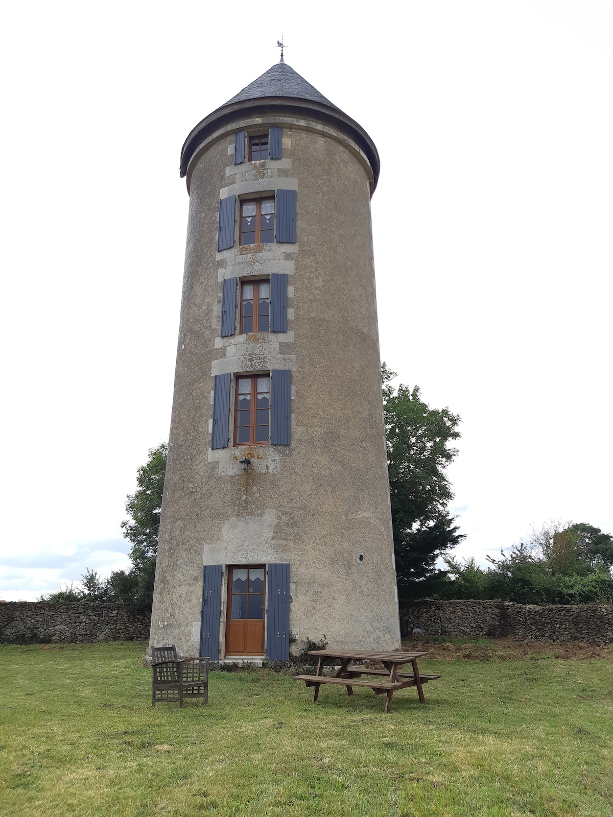 A tall, historic windmill stands with a cylindrical shape, featuring multiple windows with blue shutters. A wooden picnic table and chairs are situated in front of the structure, surrounded by an expansive grassy area and framed by a stone wall and trees in the background.