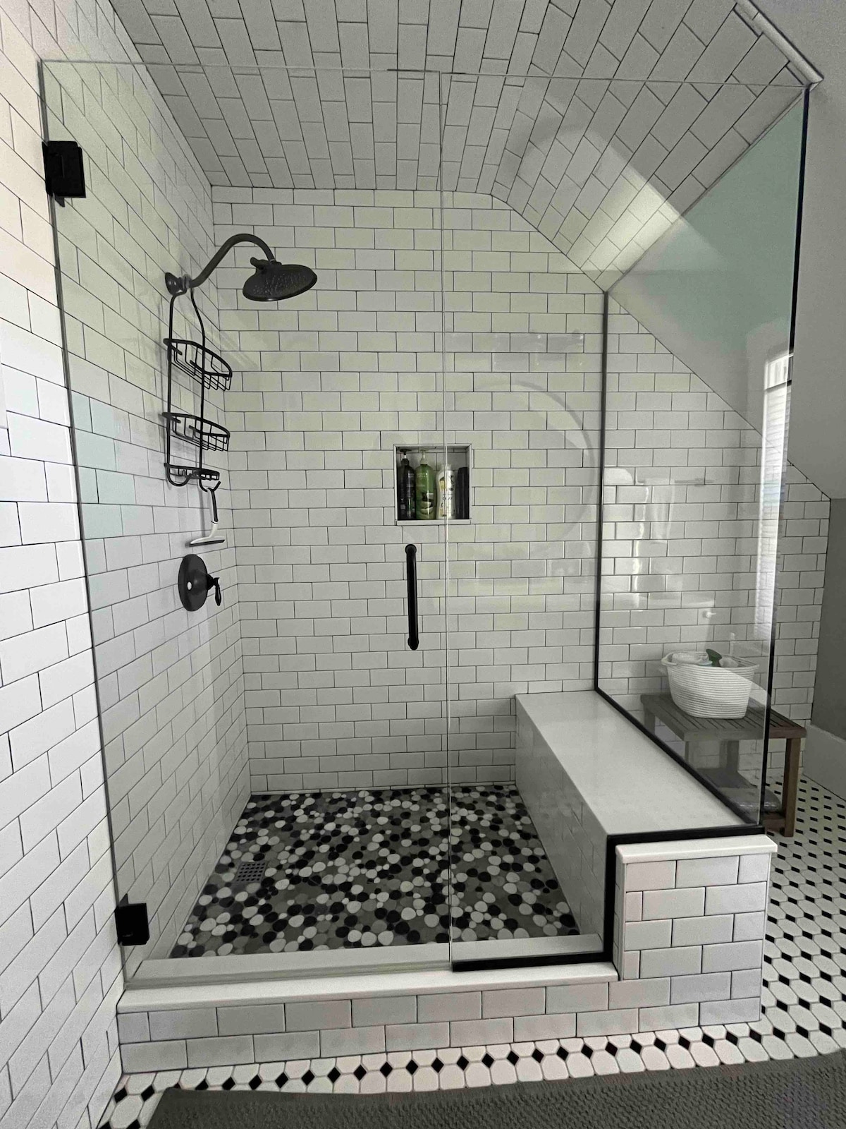 A modern shower area is showcased with white subway tiles and a decorative black and white mosaic floor. A glass enclosure allows light into the space, while a rainfall showerhead is mounted above. A bench is included for added convenience.