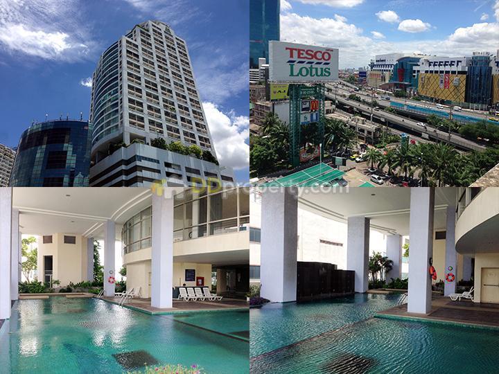 The image showcases the modern architecture of the apartment building against a clear blue sky. Views of a shopping complex and bustling streets are visible from the terrace. An inviting swimming pool, surrounded by lounge chairs, is situated in the building's outdoor area.
