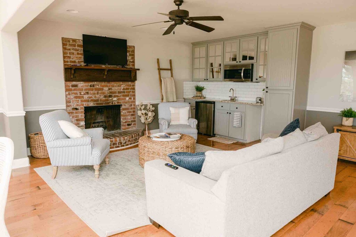 A cozy living area features a light-colored sofa and striped armchairs arranged around a round coffee table. A brick fireplace serves as a focal point, complemented by a wall-mounted television. Natural light fills the space through large windows.