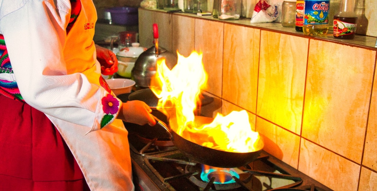 A chef is seen in a kitchen, skillfully handling a pan over a flame. Bright flames are evident, illuminating the surroundings. Various cooking ingredients are displayed on a shelf in the background, and the stove is equipped with several burners.