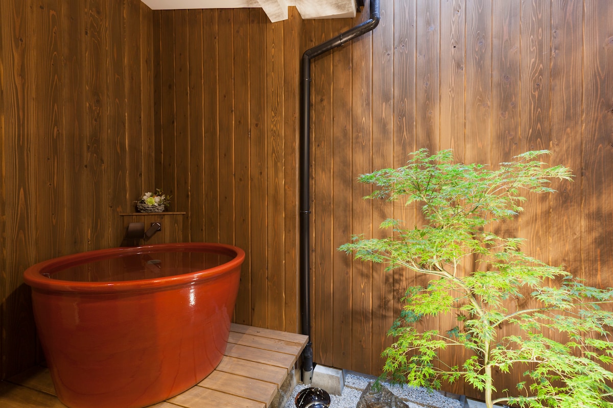 An inviting wooden space features a traditional tub, surrounded by warm-toned wooden walls. A slender green plant is positioned near the tub, enhancing the serene atmosphere. Natural light softly illuminates the area, creating a calming environment for relaxation.