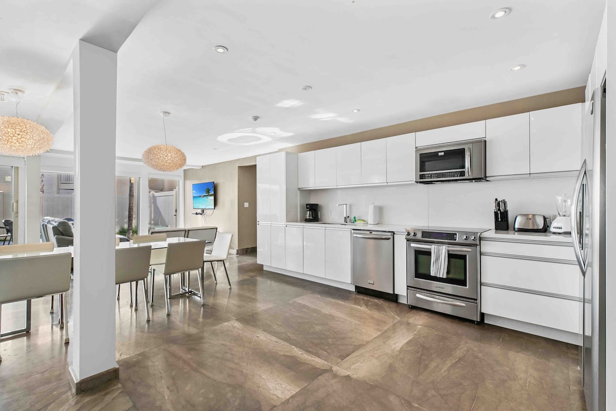 A modern kitchen is equipped with high-end stainless-steel appliances, including a stove and microwave. A dining area with seating for eight is visible, along with sleek cabinetry. Natural light fills the space through large windows, creating a bright and open atmosphere.