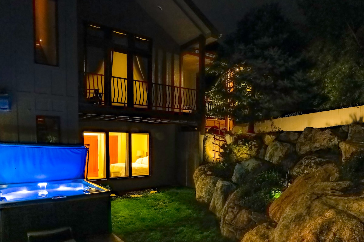 A private outdoor space showcases a luxurious hot tub with a blue cover, surrounded by natural stone features and lush grass. The building with floor-to-ceiling windows emits warm light, inviting a cozy atmosphere. Scenic trees provide a backdrop, enhancing the tranquil setting.