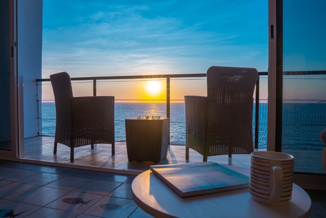 A balcony scene is captured, featuring two woven chairs and a small table overlooking the ocean. The sun sets on the horizon, casting a warm glow across the water. A coffee mug rests on a nearby table, while a magazine lies open.