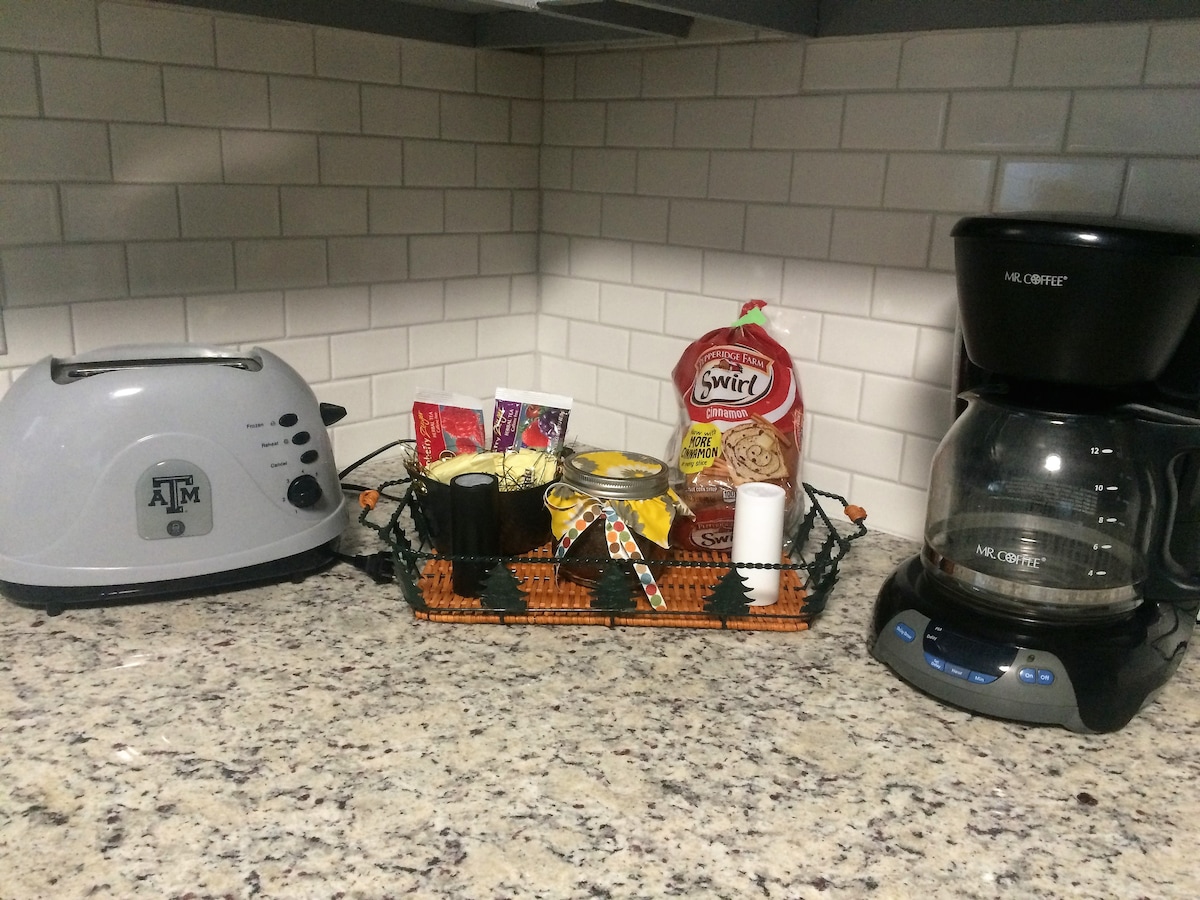 A countertop displays a toaster alongside a coffee maker. A decorative basket holds assorted coffee flavors and creamers, providing options for guests. The backdrop features a textured granite surface with white tiled backsplash, enhancing the kitchen's functionality.