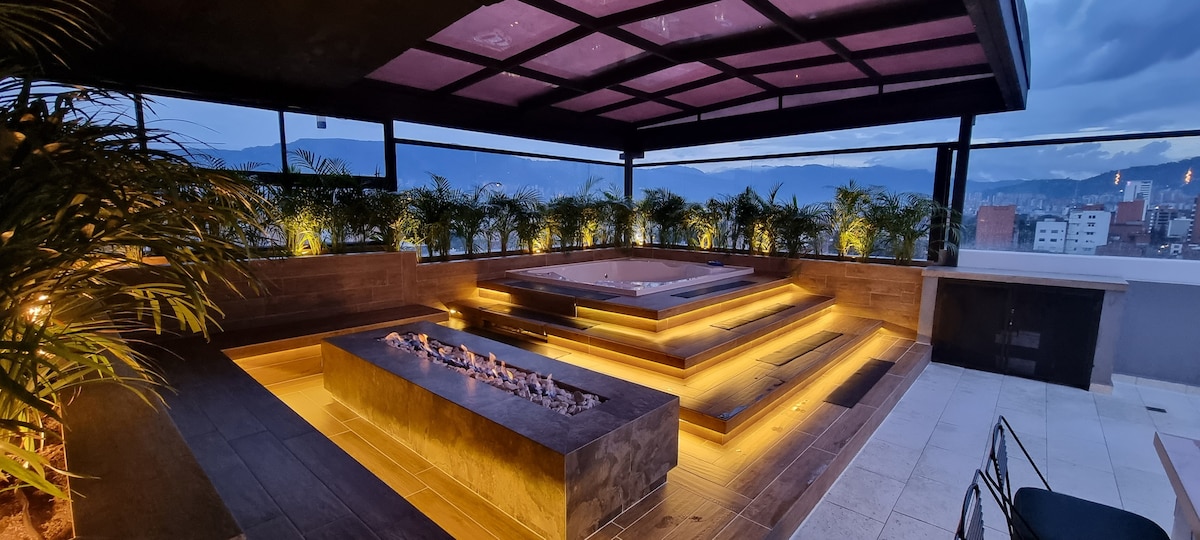 A private terrace features a spacious jacuzzi surrounded by elegant wooden decking. Soft lighting highlights the layout, creating a cozy ambiance. Lush green plants line the perimeter, adding a touch of nature, while panoramic views of the city skyline can be appreciated in the background.