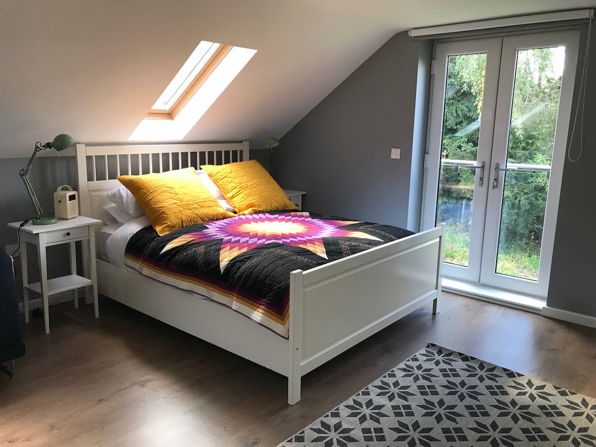 A spacious bedroom features a large bed with a colorful quilt and vibrant throw pillows. Natural light enters through a skylight and double doors that open to a view of greenery. A bedside table with a lamp and a decorative rug complete the room.