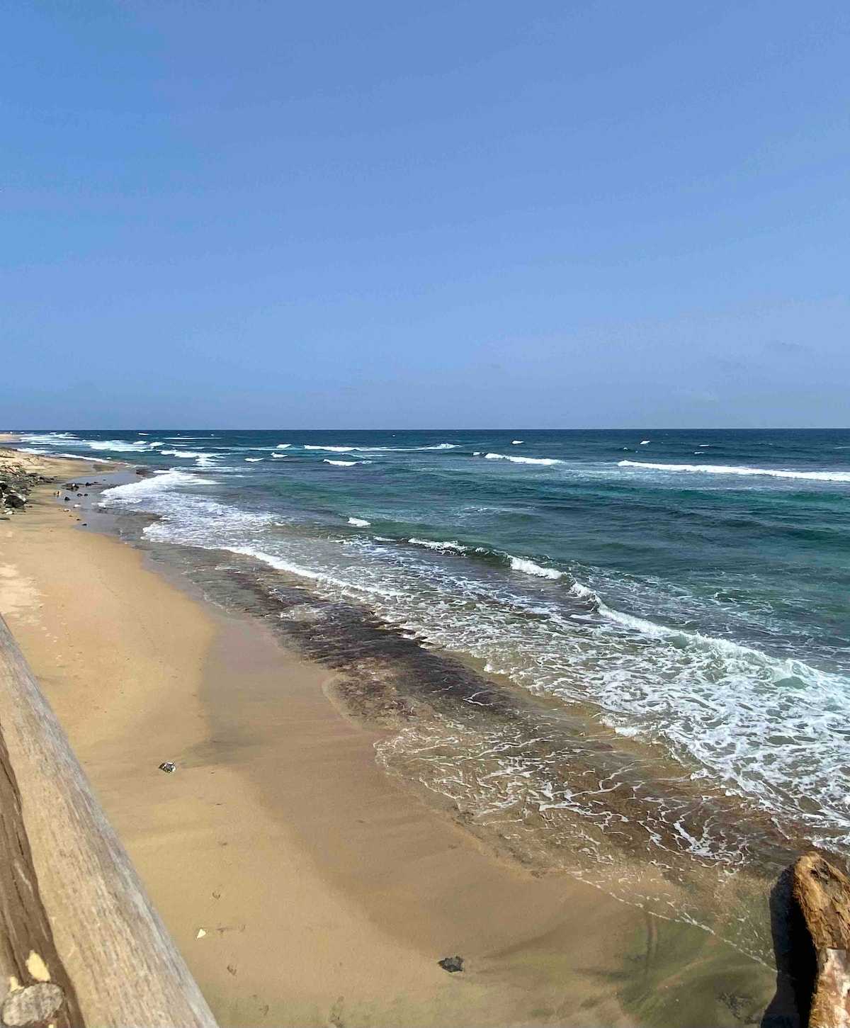 The image captures a serene beach scene, featuring gentle waves lapping against the sandy shore. Sunlight reflects off the water, while a clear blue sky stretches above. Rocky formations dot the coastline, adding to the natural beauty of the landscape.