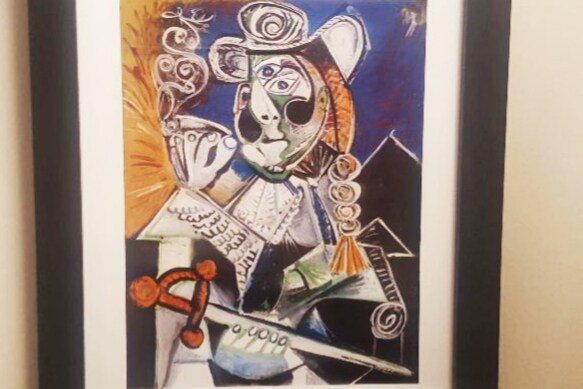 A vibrant artwork by Pablo Picasso is displayed, featuring abstract figures with distinctive features. The colors include deep blues, blacks, and splashes of orange, creating a striking visual statement. The piece is framed and hung against a neutral wall, contributing to the artistic theme of the unit.