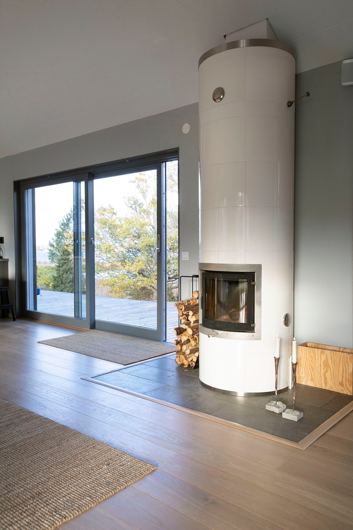A modern, cylindrical fireplace is positioned in a bright room with large glass doors leading to an outdoor terrace. The fireplace features a wood storage area and is complemented by light wooden flooring and a natural fiber rug.