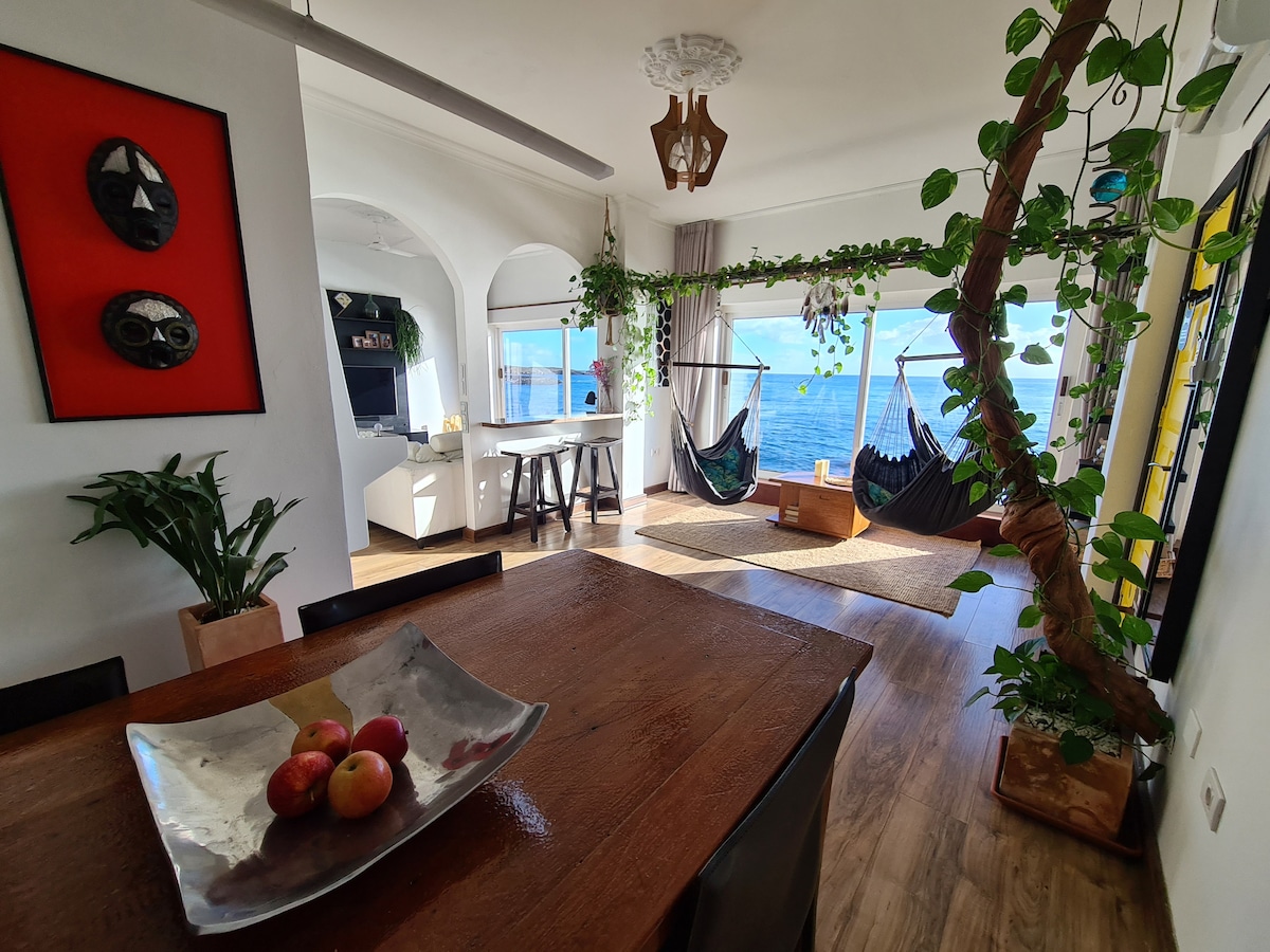 A spacious living area features a dining table with a decorative bowl of apples. Two hammocks are suspended near the large windows, offering views of the ocean. Green plants add a touch of nature, while artwork adorns the walls, creating a welcoming vibe.