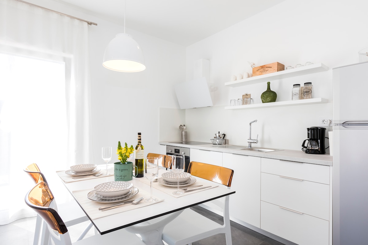 A modern kitchen area is presented with a white table set for four, featuring elegant dinnerware and a centerpiece of yellow flowers. Above, open shelves display kitchen essentials. A large window allows natural light to brighten the space, enhancing its clean and contemporary design.