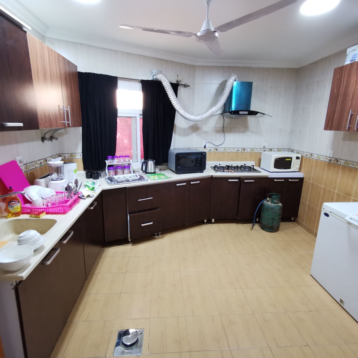 The kitchen features dark wooden cabinets and a light countertop. Appliances include a microwave, kettle, and gas stove. A fan is mounted on the ceiling, and a window provides natural light. Various kitchen items are organized on the countertop, emphasizing a functional cooking space.