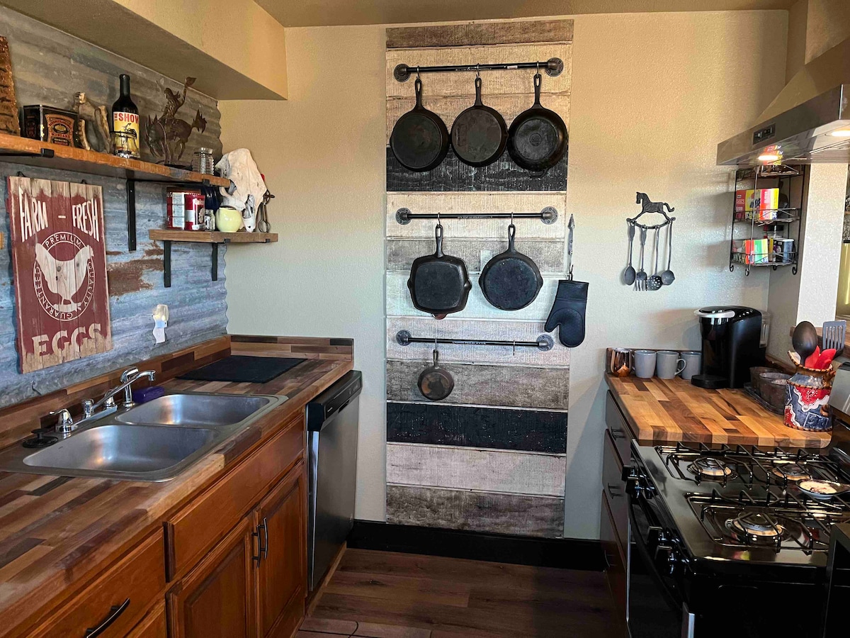 The kitchen features a rustic design with wooden cabinetry and a sleek countertop. Cast iron cookware is displayed on a wall-mounted rack, and essential appliances, including a gas stove and coffee maker, are visible. Shelves hold various kitchen items, enhancing the functional space.