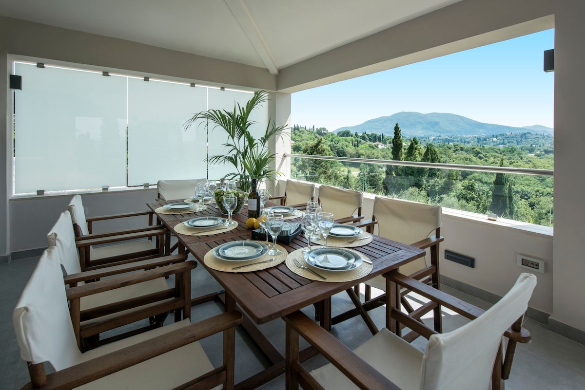 A dining area features a wooden table surrounded by eight light-colored chairs. Glassware and plates are set for a meal, with a view of lush greenery and distant hills visible through large sliding glass doors. The space is bright and airy.