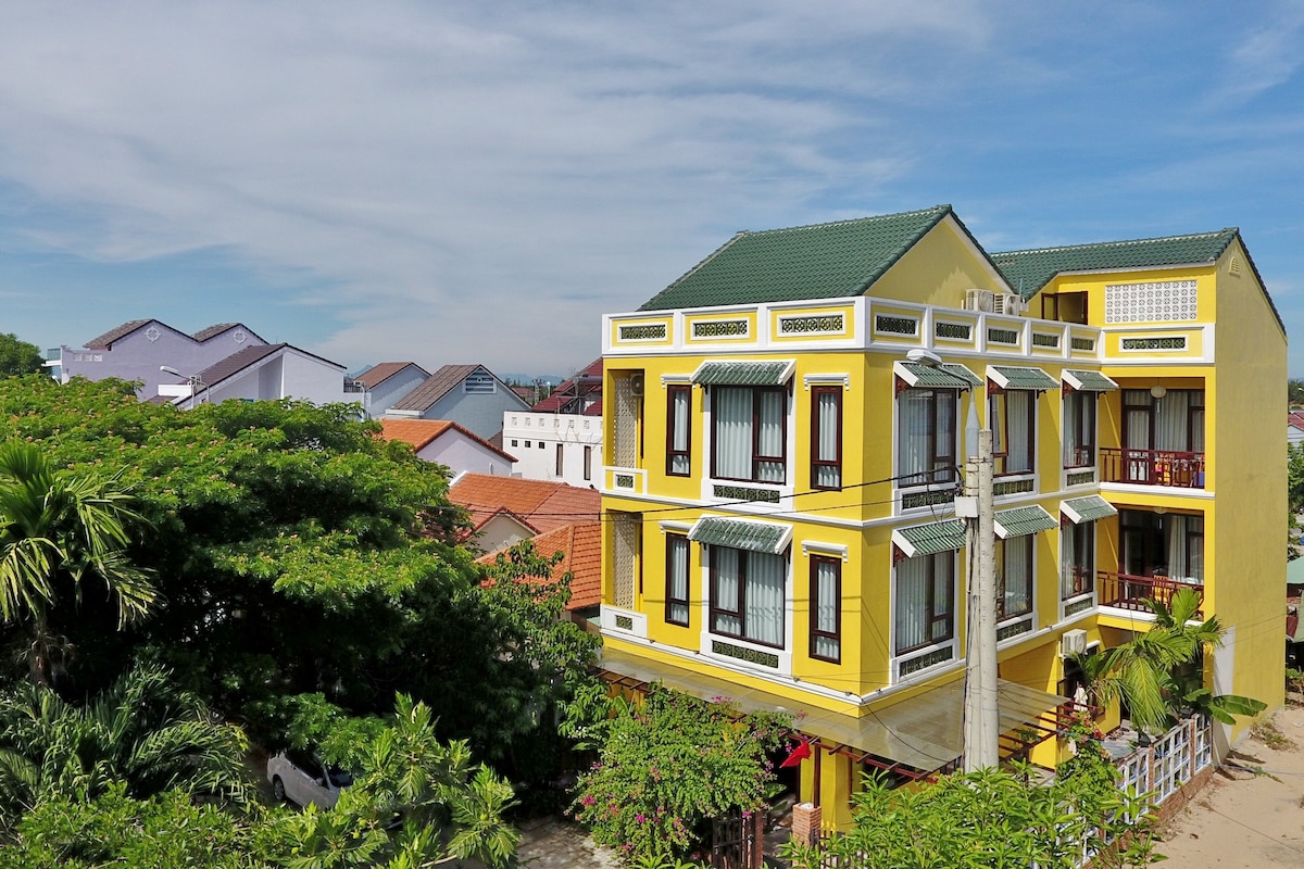 A vibrant yellow building is prominently featured, showcasing multiple balconies with large windows. Surrounding greenery and palm trees add a natural element, while the nearby rooftops create a pleasing contrast in colors. The clear blue sky enhances the overall appearance.