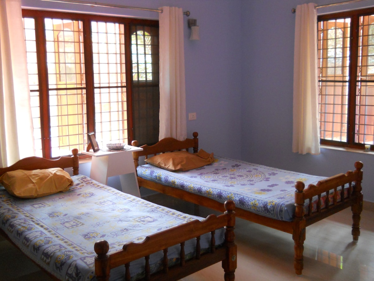 A room features two wooden beds with light blue patterned bedding and yellow pillows. Natural light enters through two large windows adorned with sheer white curtains, illuminating the neutral-toned floor and a small white table positioned between the beds.