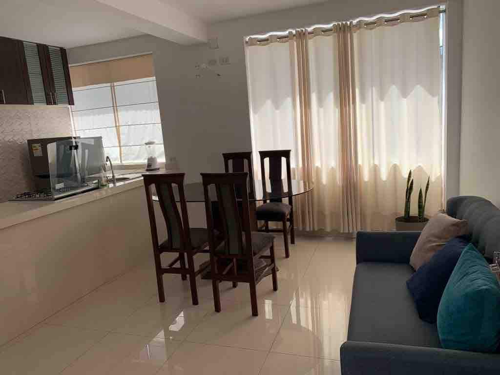 The image showcases a modern living area with a dining table for four and a comfortable sofa. Natural light enters through large windows adorned with sheer curtains, illuminating the space. A small plant is visible in the corner, adding a touch of greenery.