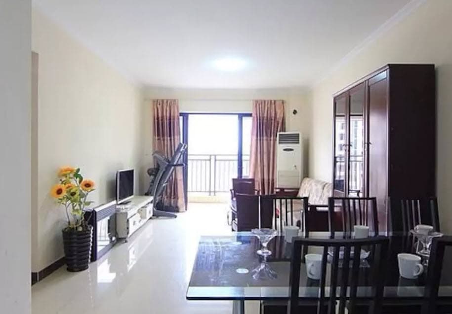 A spacious living area features large windows allowing natural light to fill the room. A dining table with chairs is present, along with a comfortable sofa. A console unit accommodates a television, while sunflowers add a touch of color to the decor.