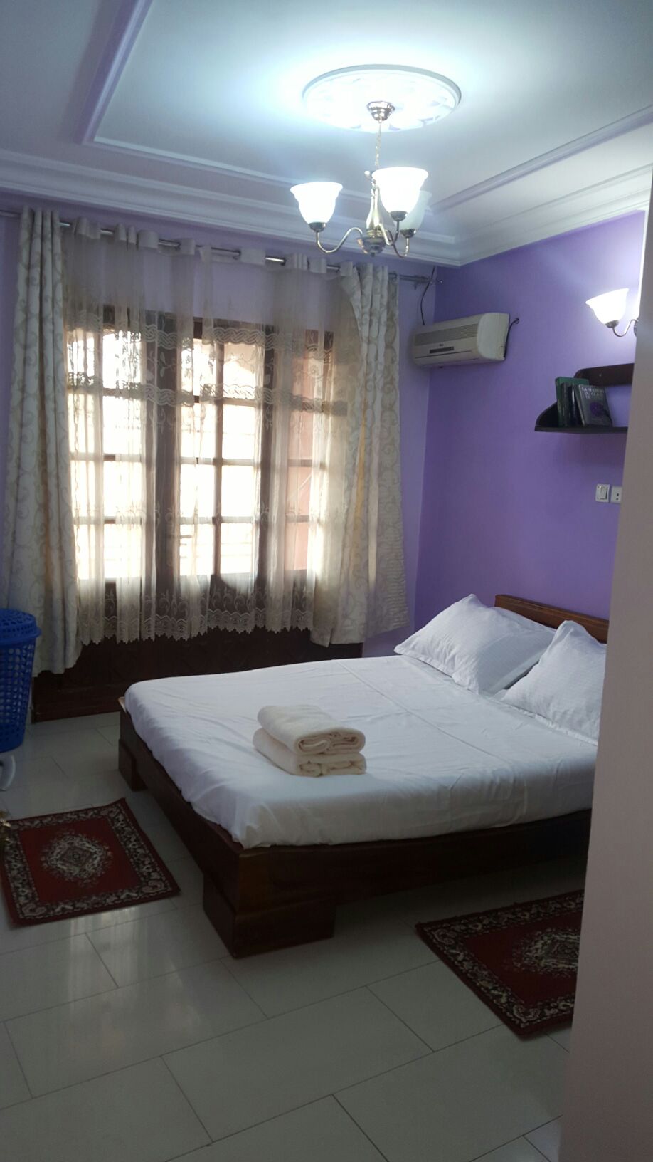 A well-lit sleeping area with lavender walls features a spacious bed, neatly made with white linens. Two towels are neatly stacked at the foot of the bed. Natural light filters through sheer curtains, illuminating the room. A small shelf is positioned above the bed.