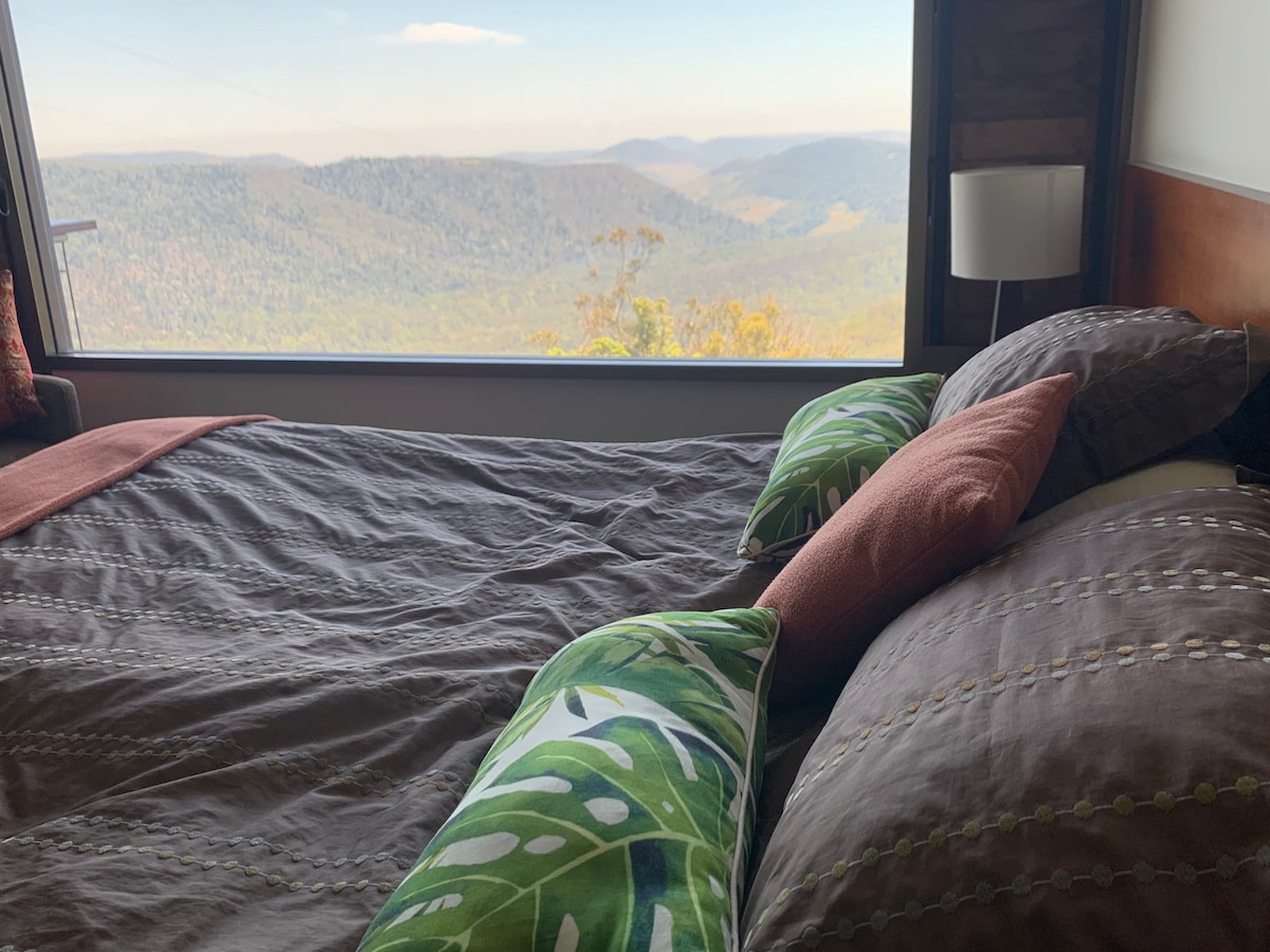 A comfortable sleeping area is presented with a spacious bed adorned with patterned bedding and colorful pillows. A large window captures expansive views of the surrounding mountains and valleys, allowing natural light to fill the space.