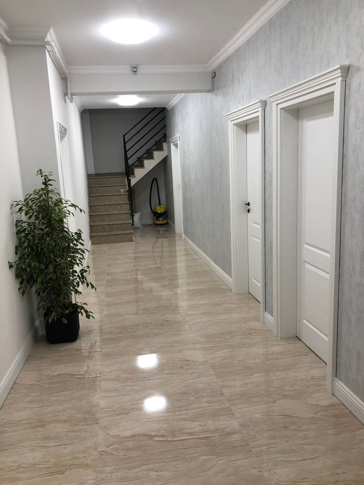 A well-lit hallway showcases smooth, glossy tiles and light-colored walls. Two closed doors are visible on one side, with a small potted plant positioned near the end. A staircase leads to an upper level, enhancing the sense of space.