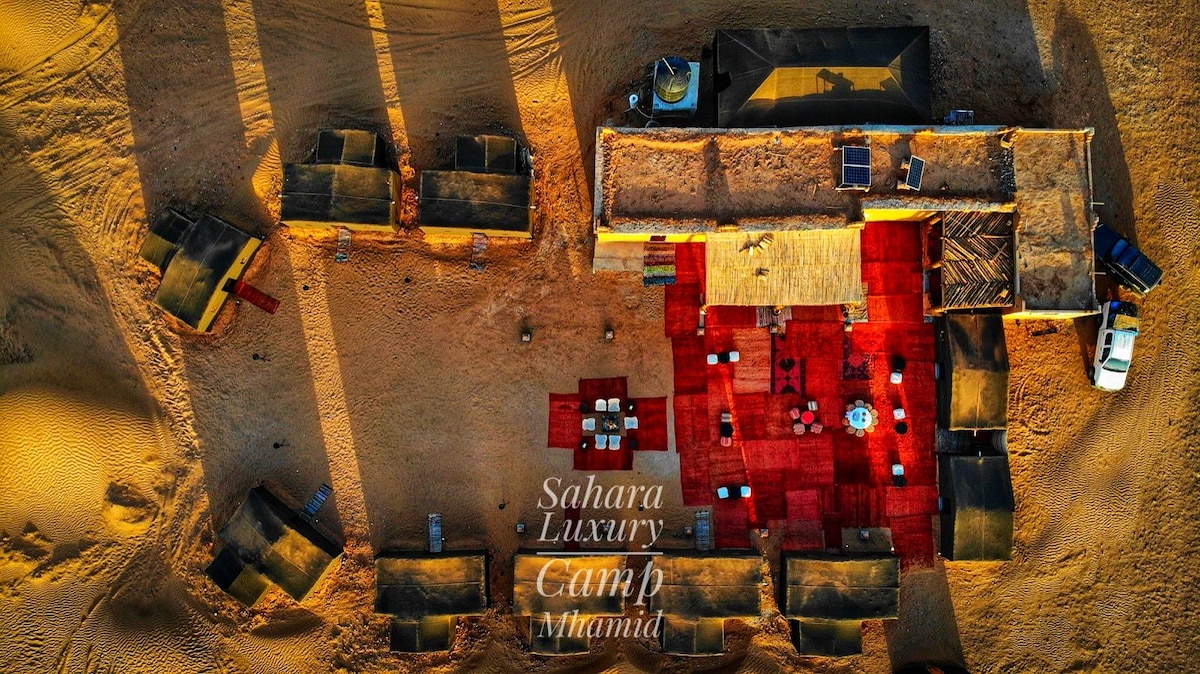 An aerial view captures the layout of Sahara Luxury Camp M'hamid, featuring several luxury tents arranged around a central courtyard. A vibrant red carpet is spread across the communal area, contrasting with the desert landscape, while shaded seating areas are visible under fabric canopies.
