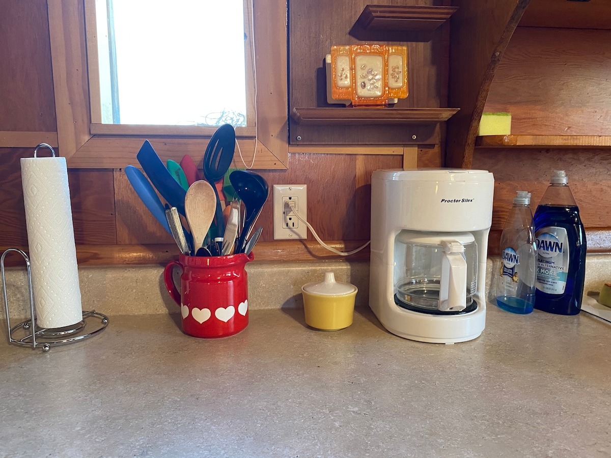 A kitchen counter is displayed with essential items. A bright red container holds various cooking utensils. A small coffee maker is positioned beside a yellow dish, while bottles of dish soap and a paper towel roll are neatly arranged on the surface. Natural light enters through a nearby window.
