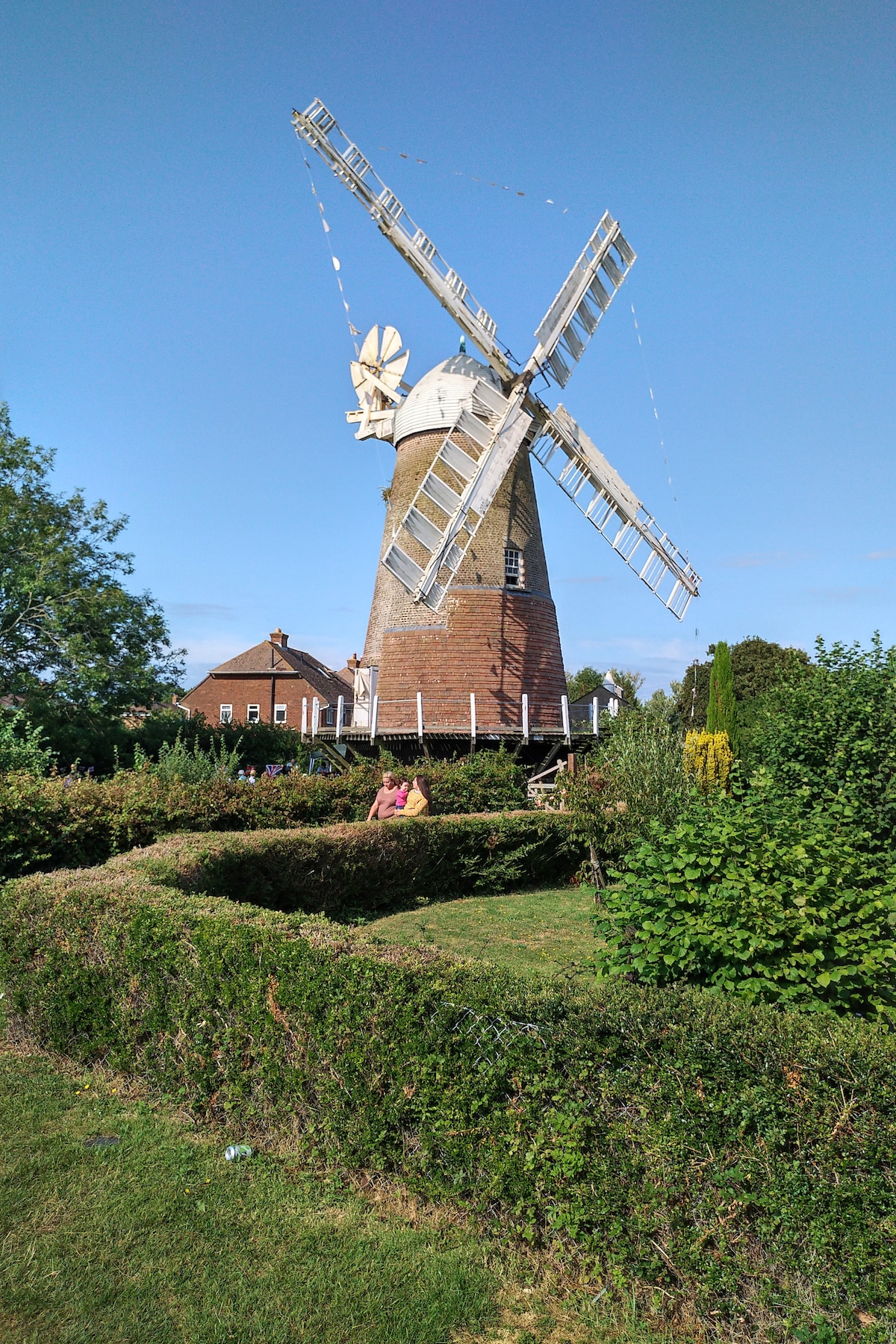 A historic windmill stands prominently against a clear blue sky, surrounded by lush greenery. The windmill's sails are visible, rotating gently in the breeze. People can be seen enjoying the space nearby, framed by well-maintained hedges and grass.