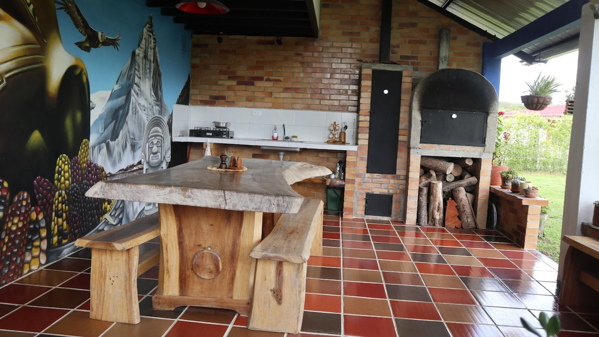 An organic gourmet kitchen area features a large wooden table surrounded by rustic benches. The space is decorated with a colorful mural and includes a traditional oven with stacked firewood nearby. Natural light fills the area through overhead windows.