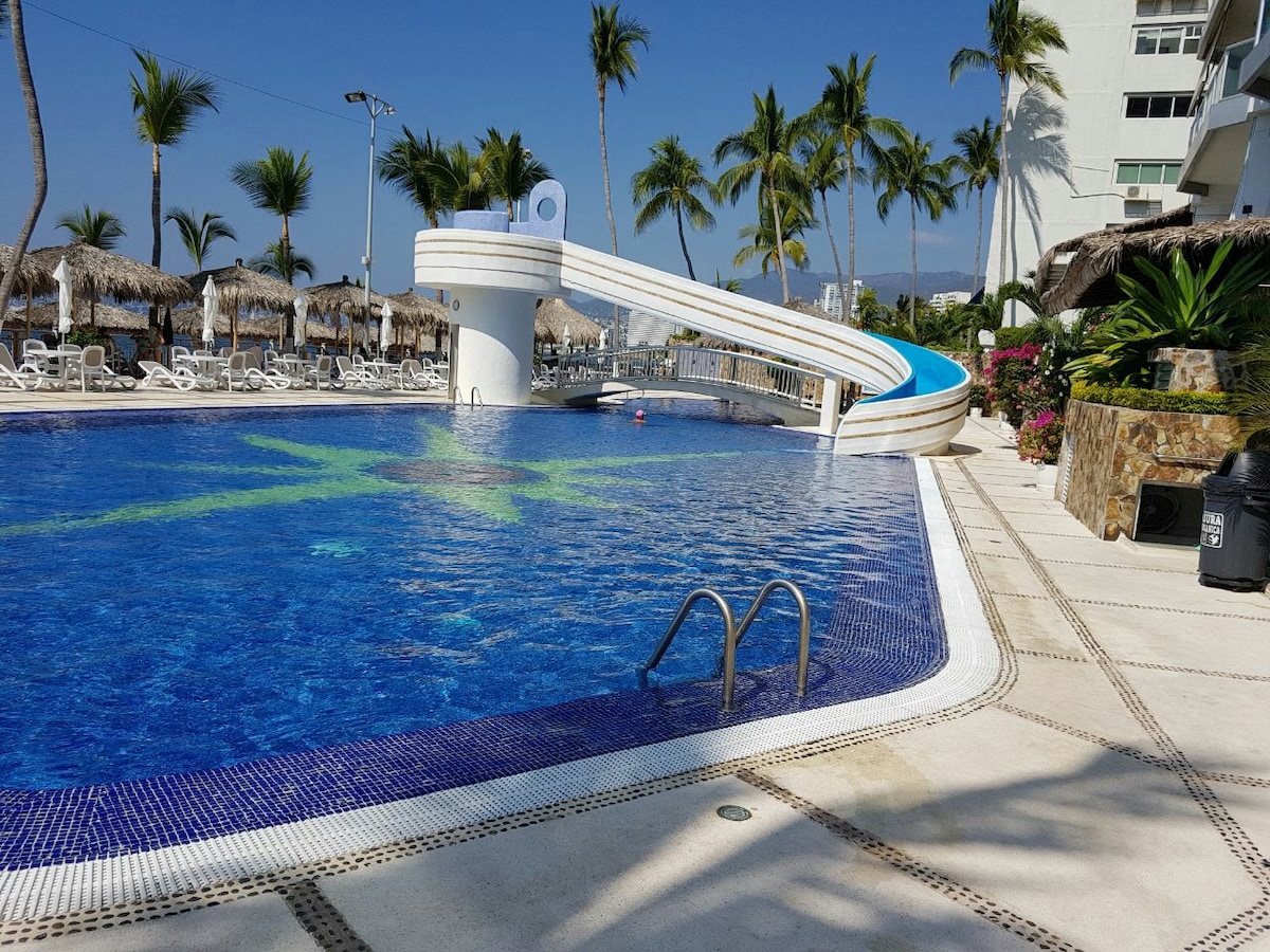 A large swimming pool is depicted, featuring a water slide that curves gracefully into the water. Surrounding the pool, palm trees stand tall against a clear blue sky. Lounge chairs are positioned nearby, and vibrant flowers add a touch of color to the scene.