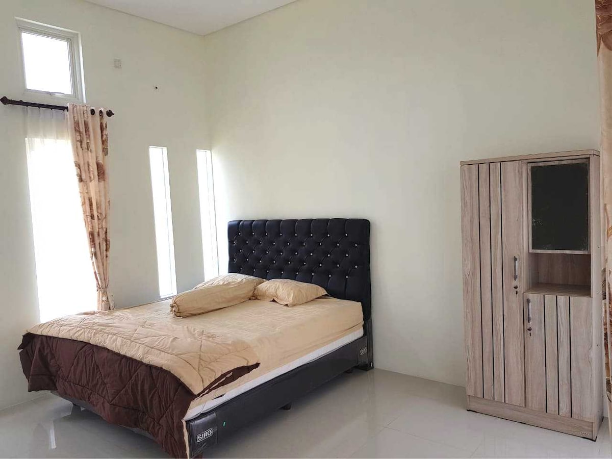 A spacious bedroom features a dark upholstered bed with an elegant headboard, complemented by a light brown blanket. Natural light filters through two narrow windows adorned with sheer curtains, alongside a wooden wardrobe with a simple design.