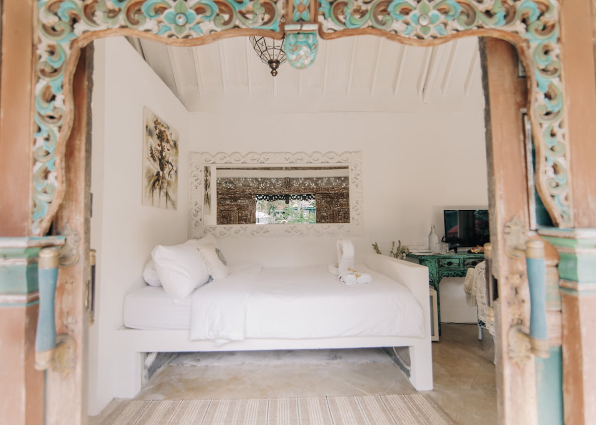 The entrance features intricate wooden doors that open to reveal a spacious room with a comfortable bed adorned with white linens. A mirror reflects natural light, enhancing the airy atmosphere. A decorative desk complements the minimalist design, inviting relaxation.