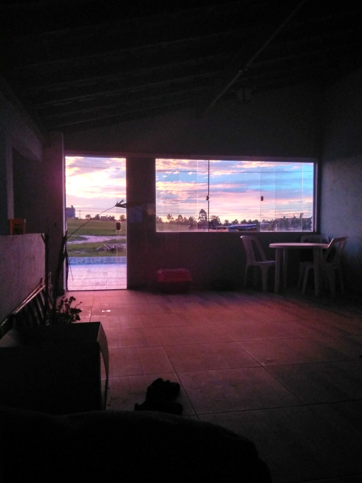 An inviting interior space is shown with large windows offering a view of a colorful sunset. The room features tiled flooring and a table with chairs positioned nearby. A glimpse of the outdoor landscape is visible through the glass, enhancing the ambiance.