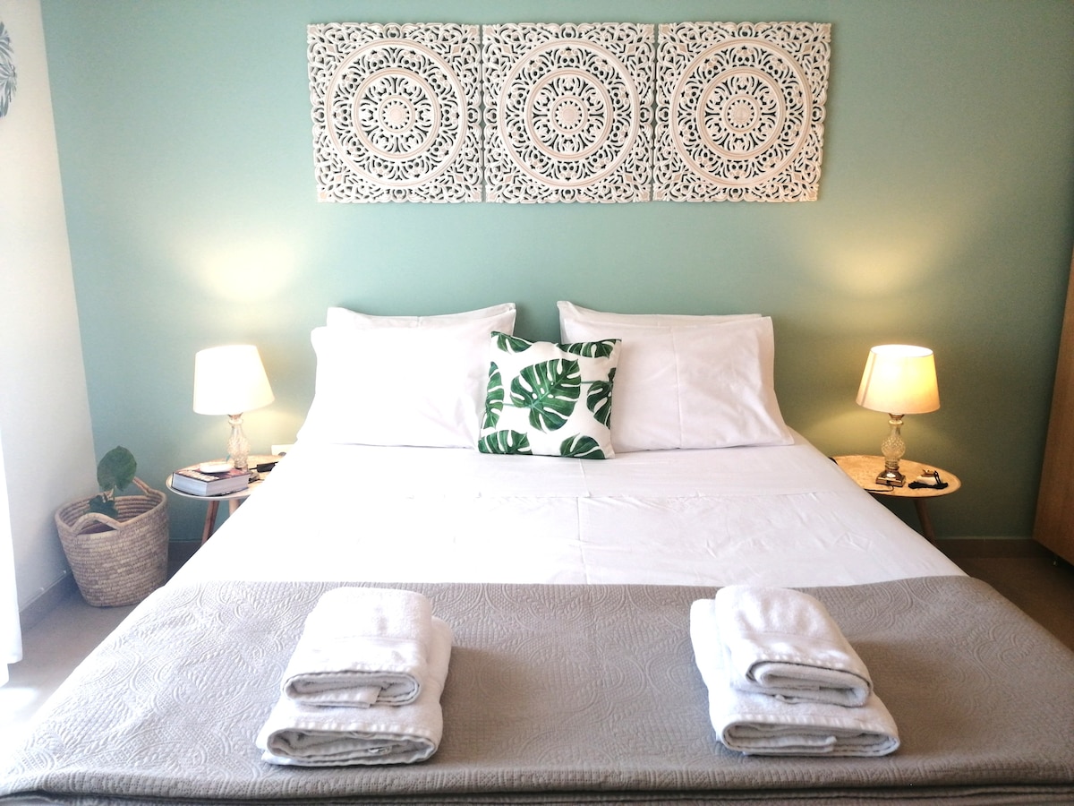 A spacious bed is centered against a soft green wall, adorned with three decorative wall hangings. Two bedside lamps provide gentle illumination, with a small plant and a decorative pillow featuring a leaf pattern adding subtle touches of nature and comfort.