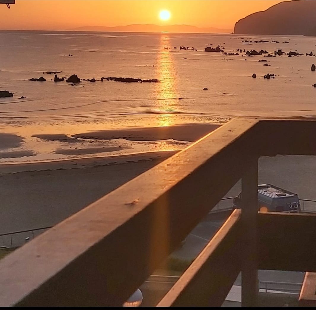 A sunset view over the calm sea is captured from a balcony, with warm orange and yellow hues reflecting on the water. The shoreline is visible, accompanied by distant rock formations, while a gentle light creates a serene ambiance.