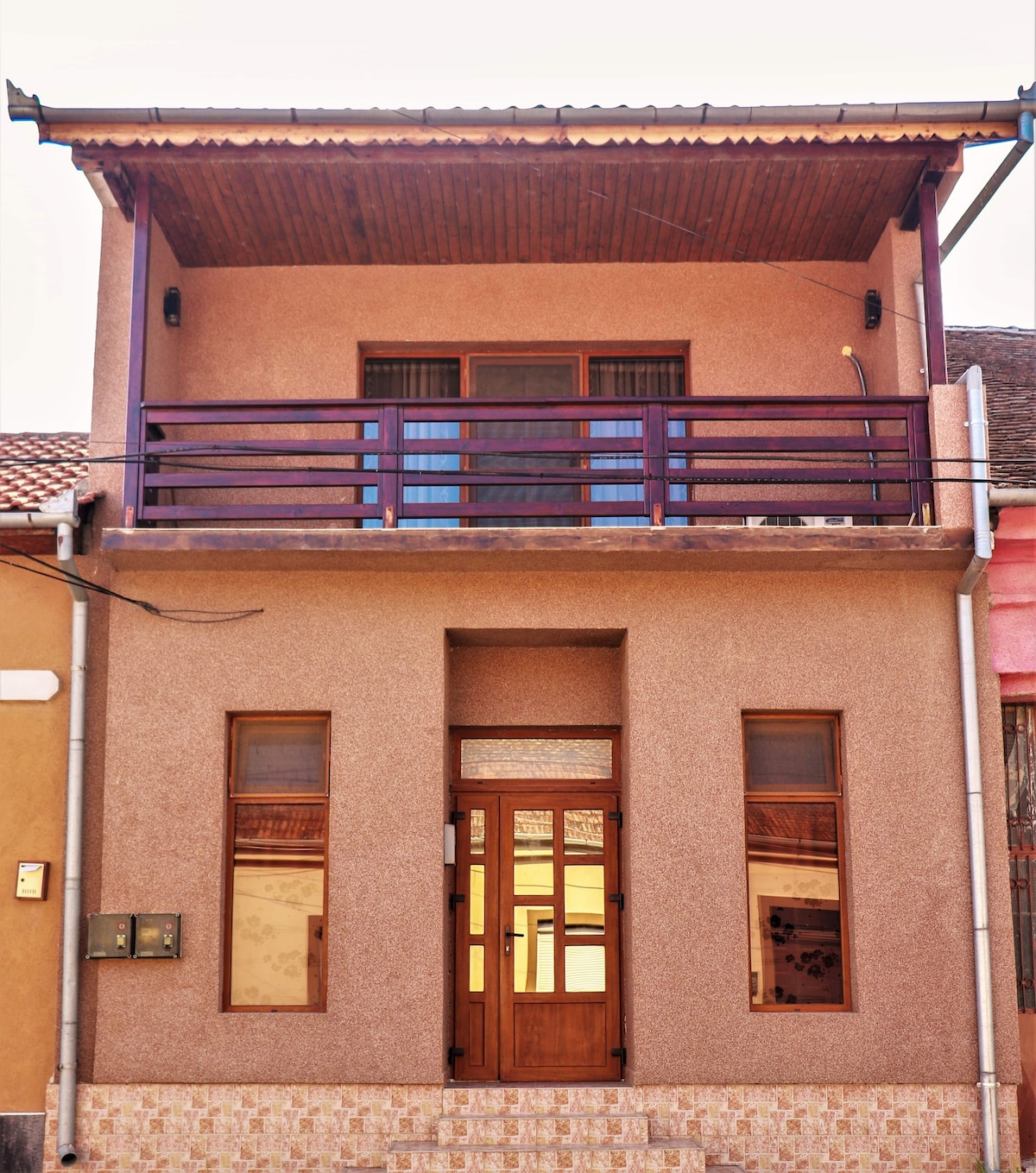 The exterior of a two-story building features a textured beige facade and a layered porch supported by dark columns. A wooden front door is flanked by tall windows, providing natural light. A balcony with a railing extends above, offering a view of the surrounding area.
