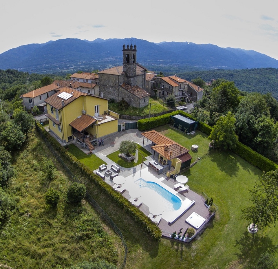 An aerial view captures the luxurious villa surrounded by lush greenery, with a private, inviting pool at the center. The rustic charm of the nearby village and its tall clock tower enhance the tranquil setting against the backdrop of rolling hills.