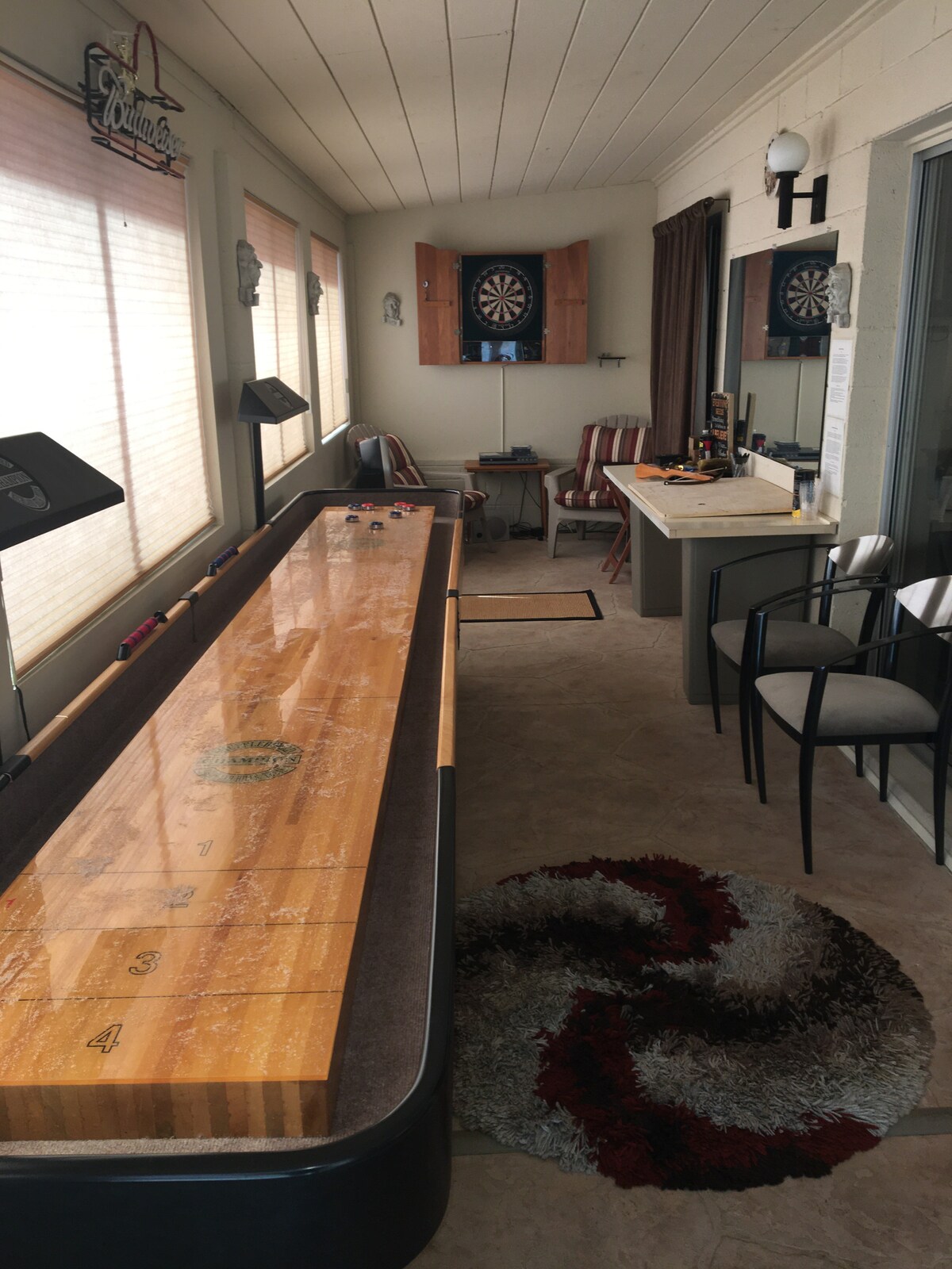 A game room is equipped with a shuffleboard table and electronic dartboard. Natural light enters through large windows, complemented by light-colored walls. A small bar area with stools is visible, alongside a patterned area rug that adds a touch of texture to the space.
